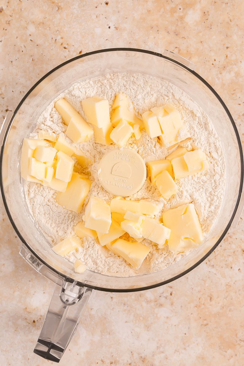 A food processor bowl filled with flour and cubes of butter, ready to be mixed, sits on a beige countertop.