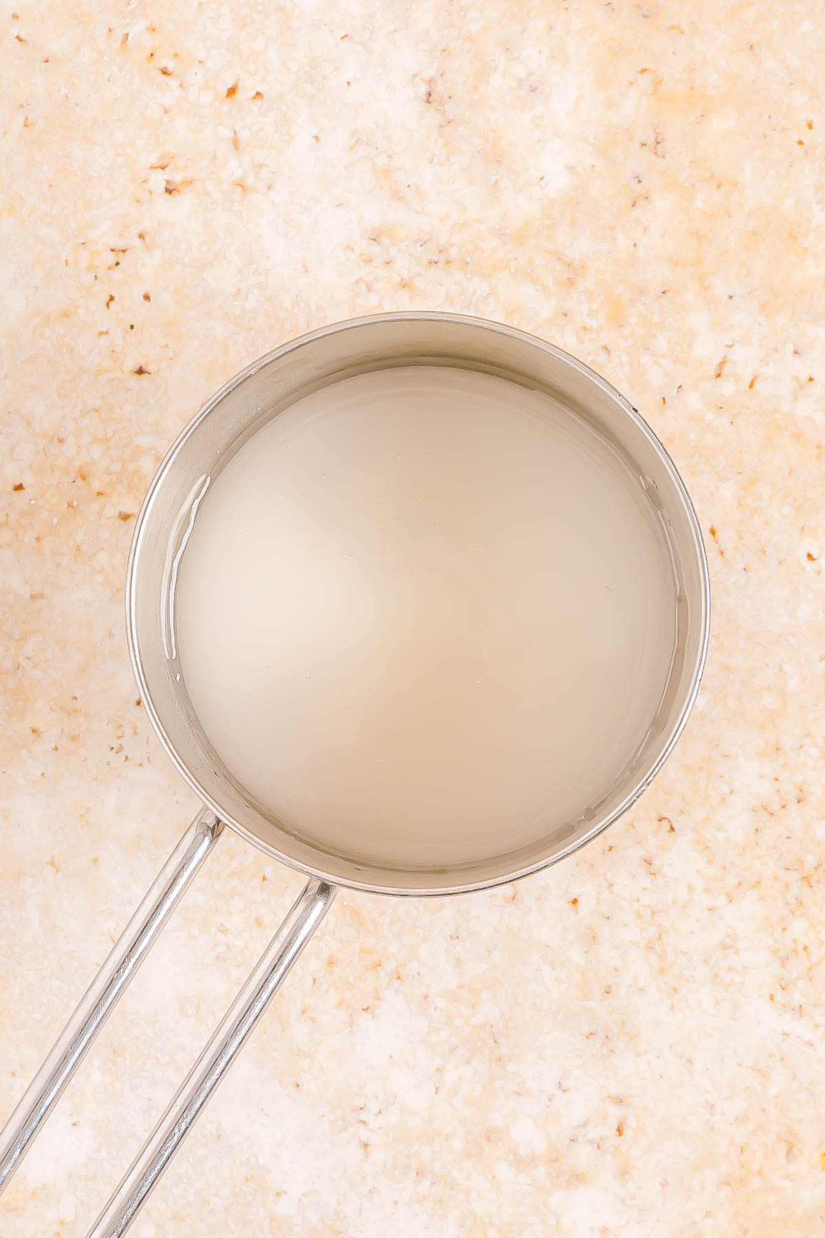 A metal saucepan filled with a clear liquid is placed on a light beige, speckled surface.