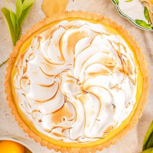 A lemon meringue pie with golden-brown swirled meringue topping on a light surface, surrounded by lemons, green leaves, and a decorative plate with gold forks.