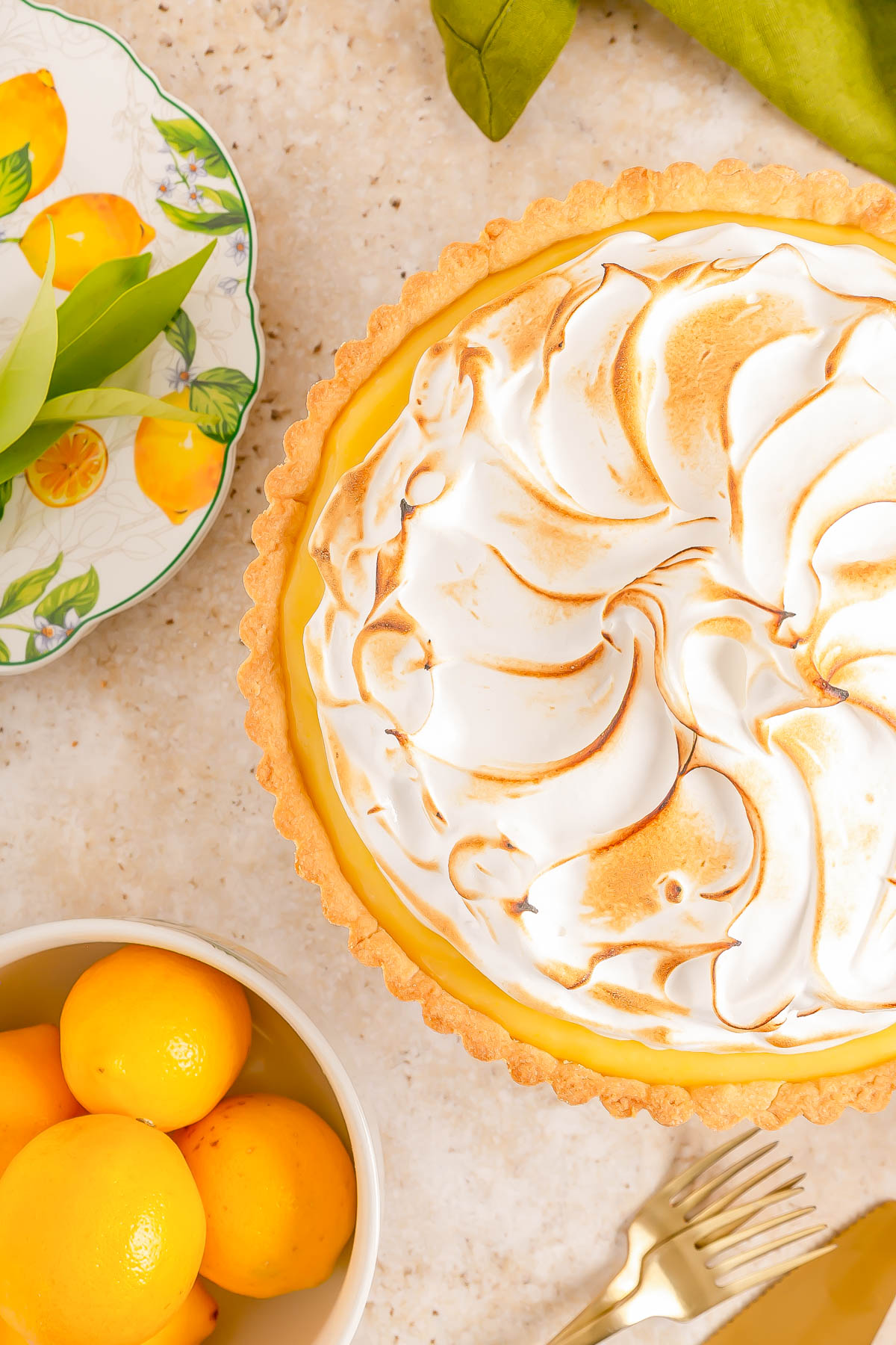 A lemon meringue pie with toasted meringue topping sits on a countertop next to a bowl of lemons, a plate with lemon designs, and two gold forks.