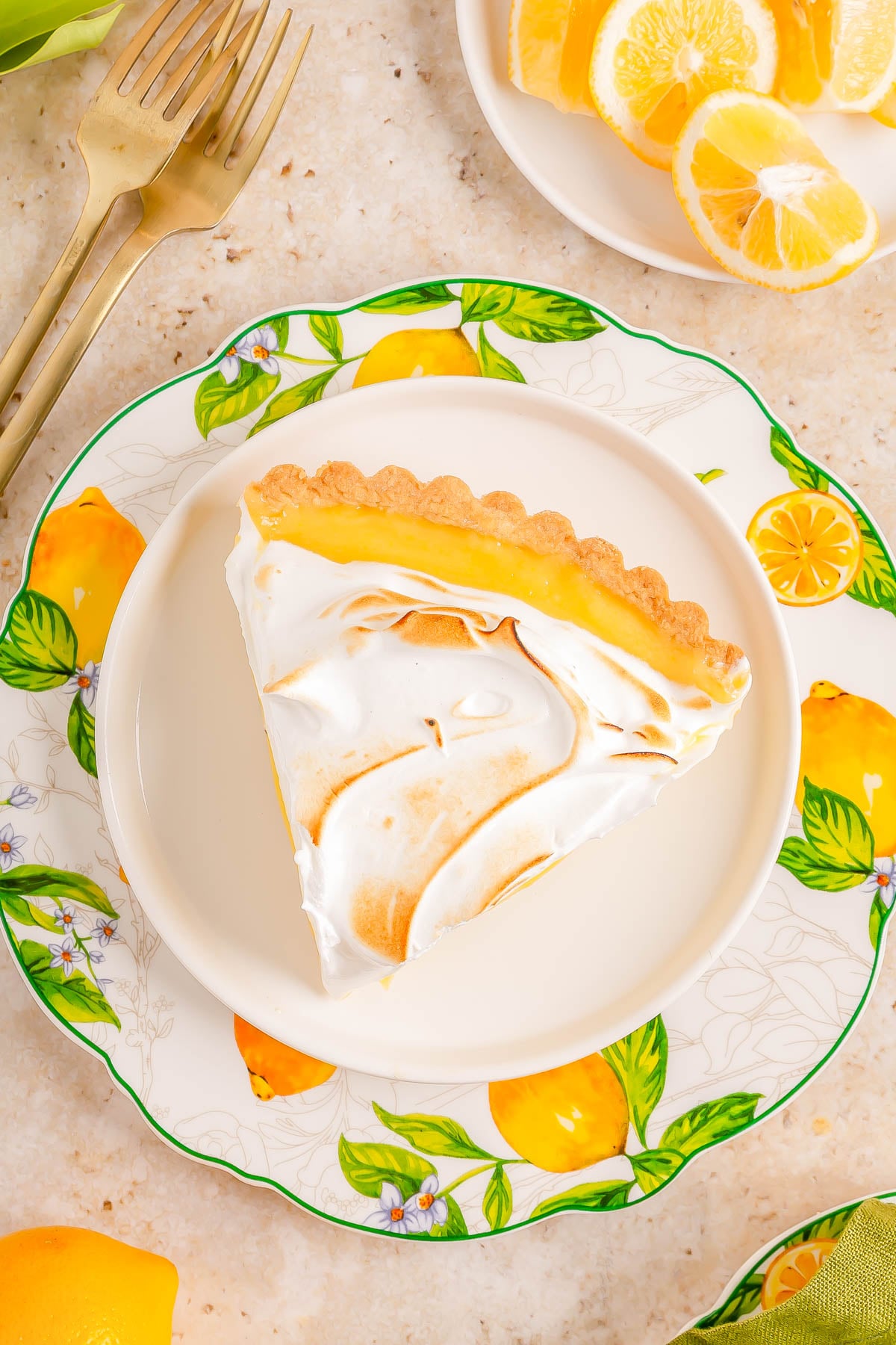 A slice of lemon meringue pie on a white plate with a lemon pattern, next to lemon slices and gold forks on a light countertop.