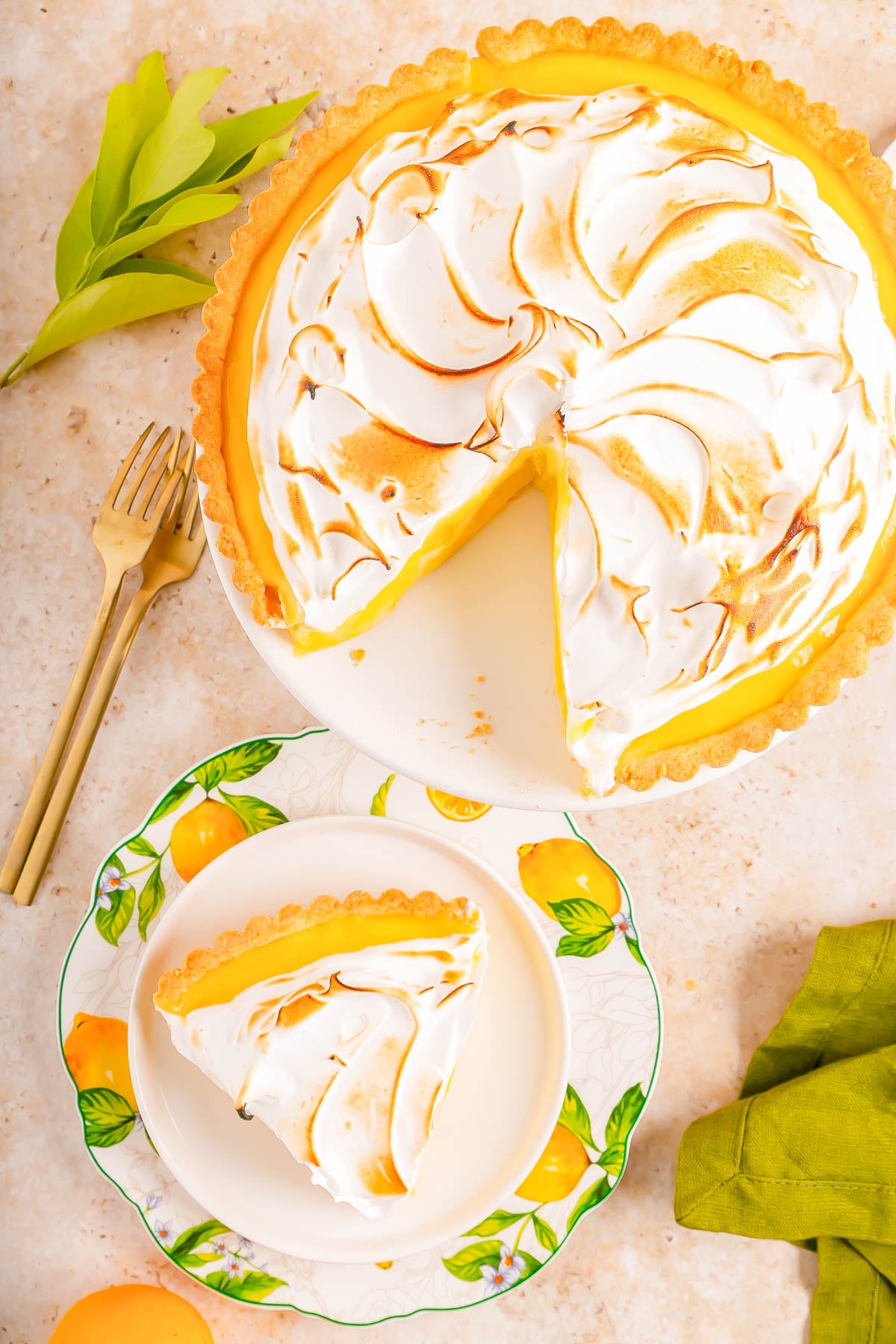 A lemon meringue pie with one slice served on a decorative plate, accompanied by a fork and green napkin on a light-colored surface.