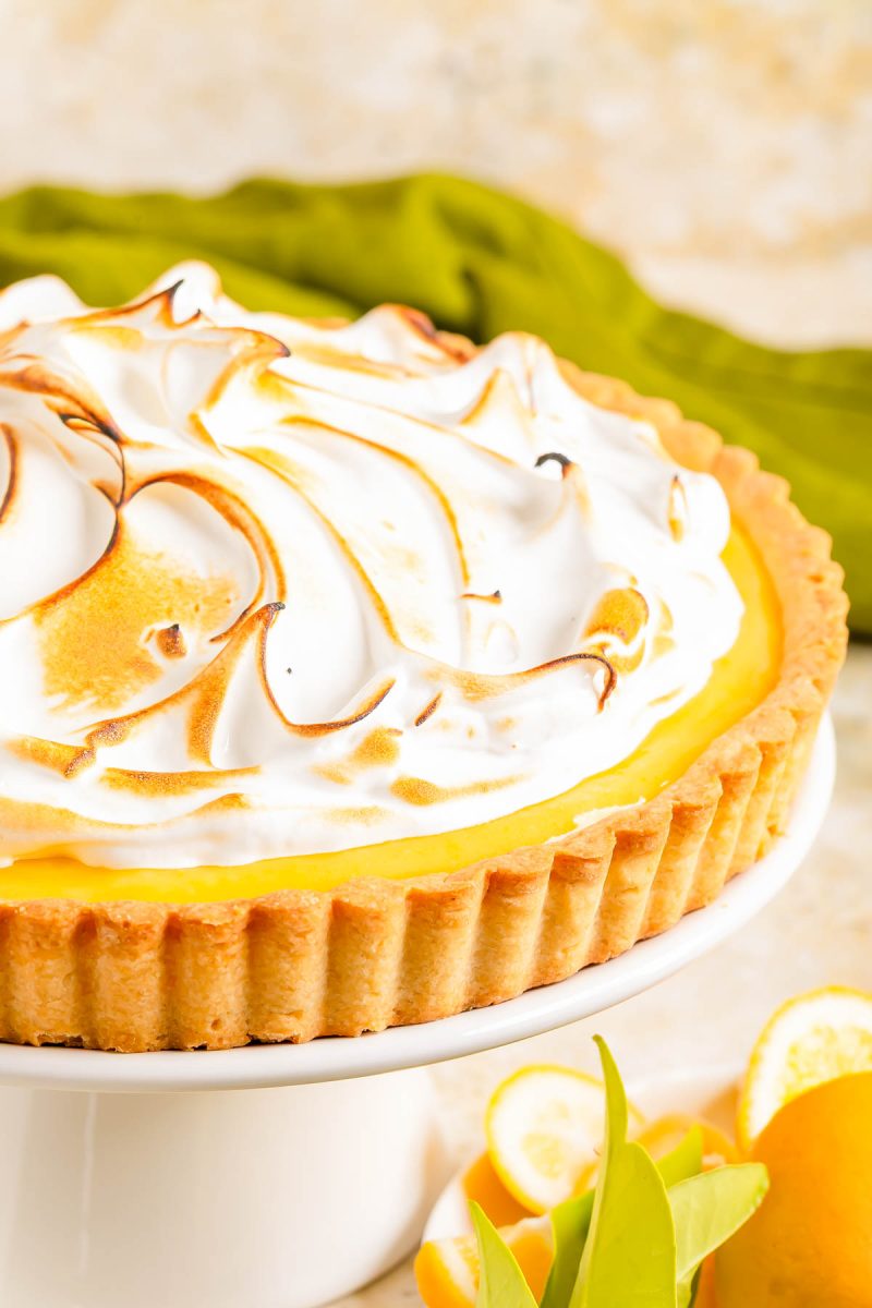 A lemon meringue tart with toasted meringue topping sits on a white cake stand, with sliced lemons and green leaves beside it.