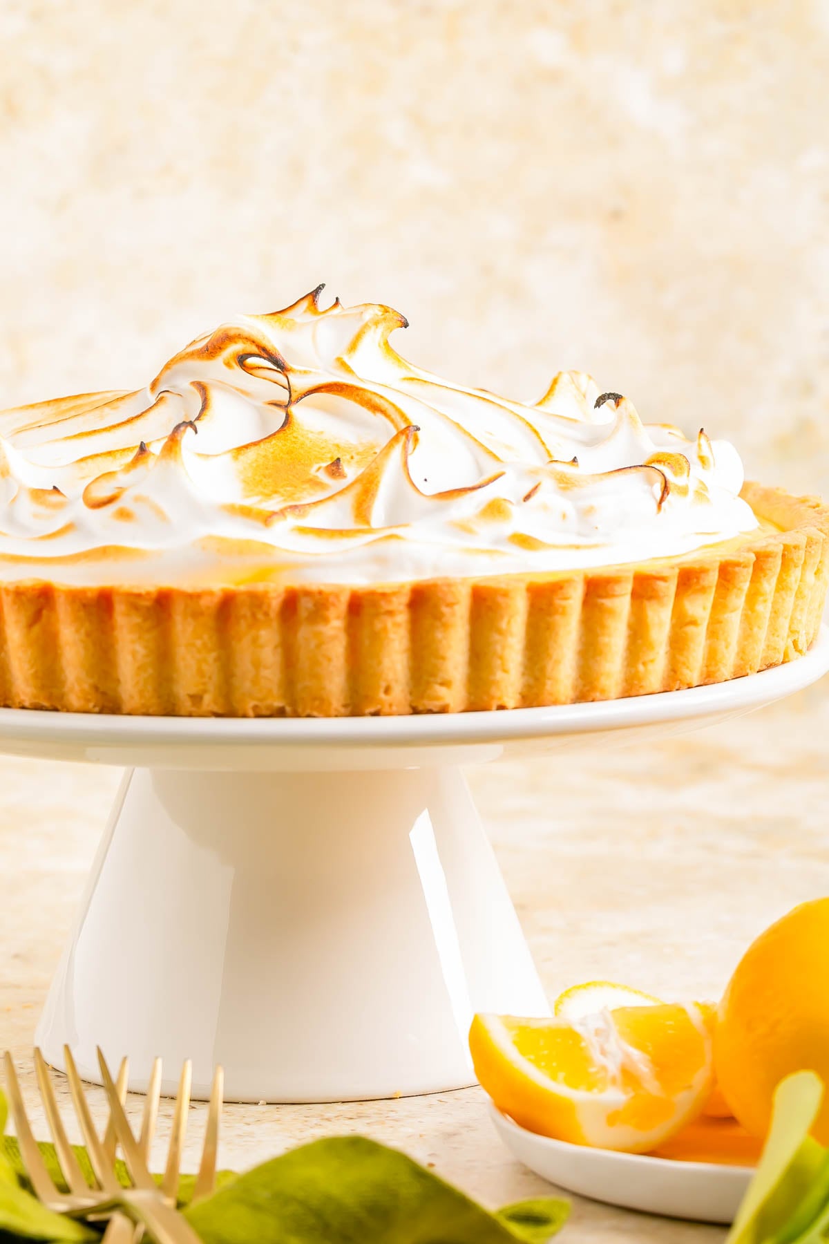 A lemon meringue pie with toasted peaks sits on a white cake stand; lemon slices and a fork are placed beside it.