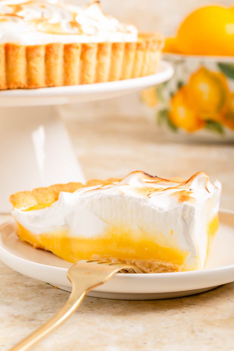 A slice of lemon meringue pie on a white plate with a gold fork, with the rest of the pie and lemons in the background.