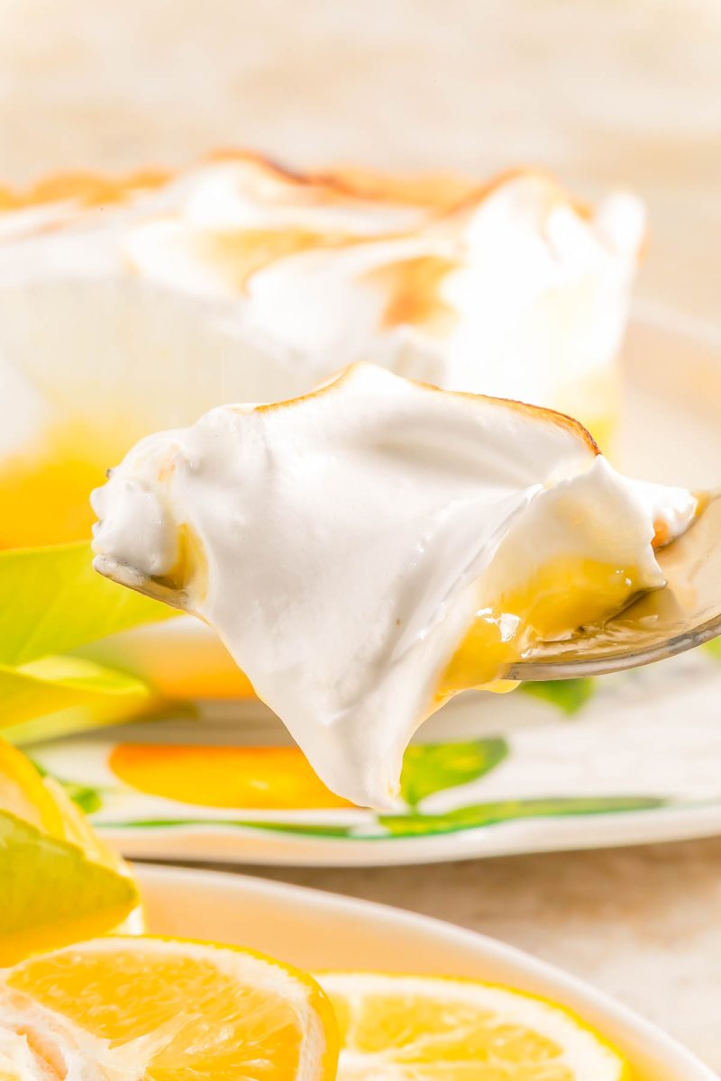 A close-up of a spoonful of lemon meringue pie showing a layer of lemon filling and fluffy meringue, with lemon slices and a pie in the background.