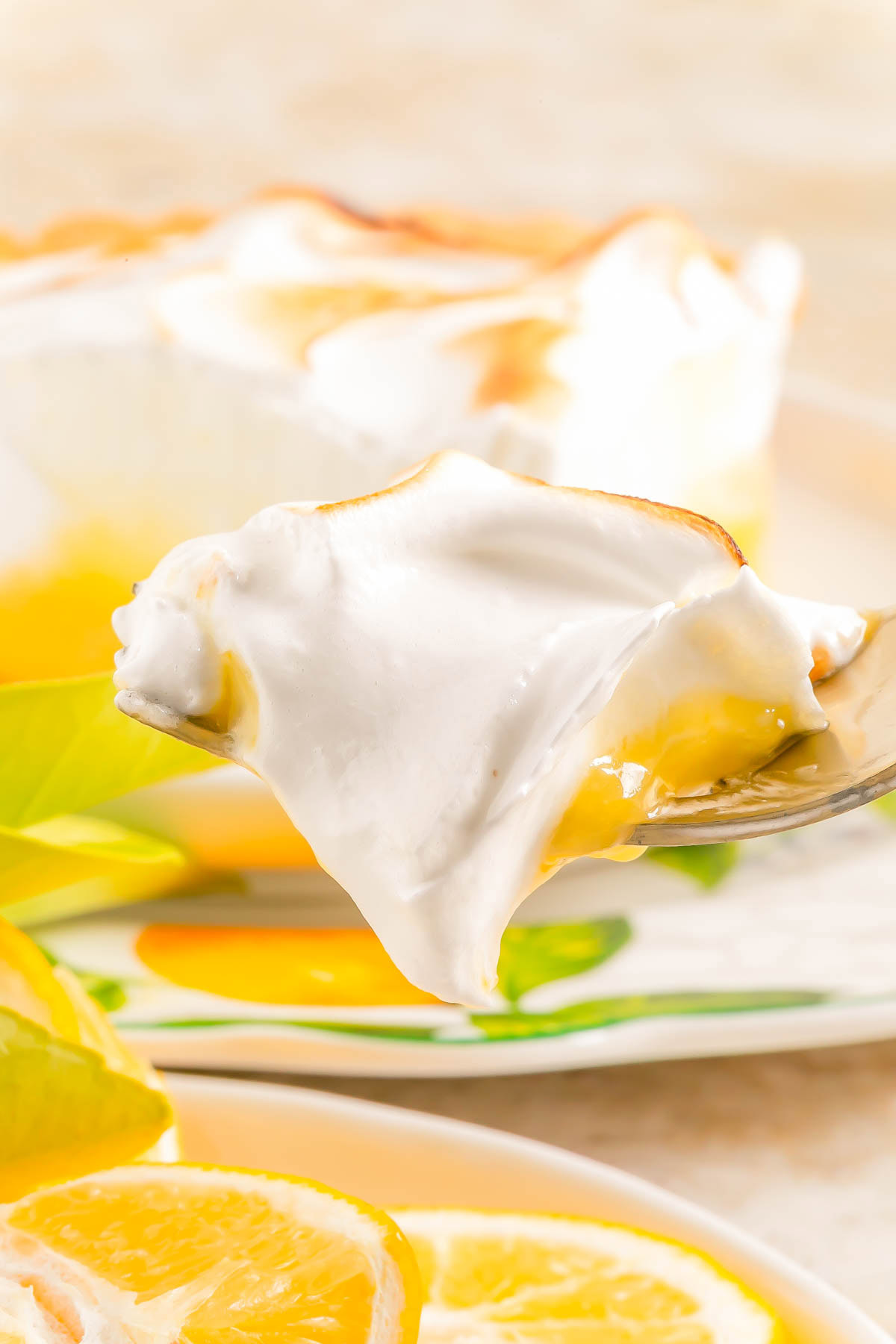 A close-up of a spoonful of lemon meringue pie showing a layer of lemon filling and fluffy meringue, with lemon slices and a pie in the background.