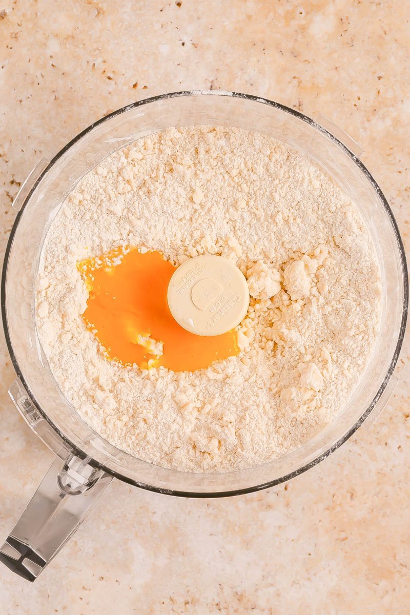 A food processor bowl contains flour, butter, and a cracked egg on top, ready to be mixed for dough preparation.