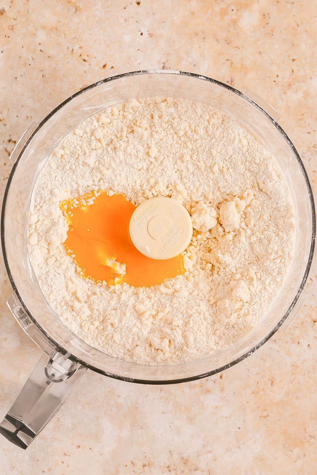 A food processor bowl contains flour, butter, and a cracked egg on top, ready to be mixed for dough preparation.