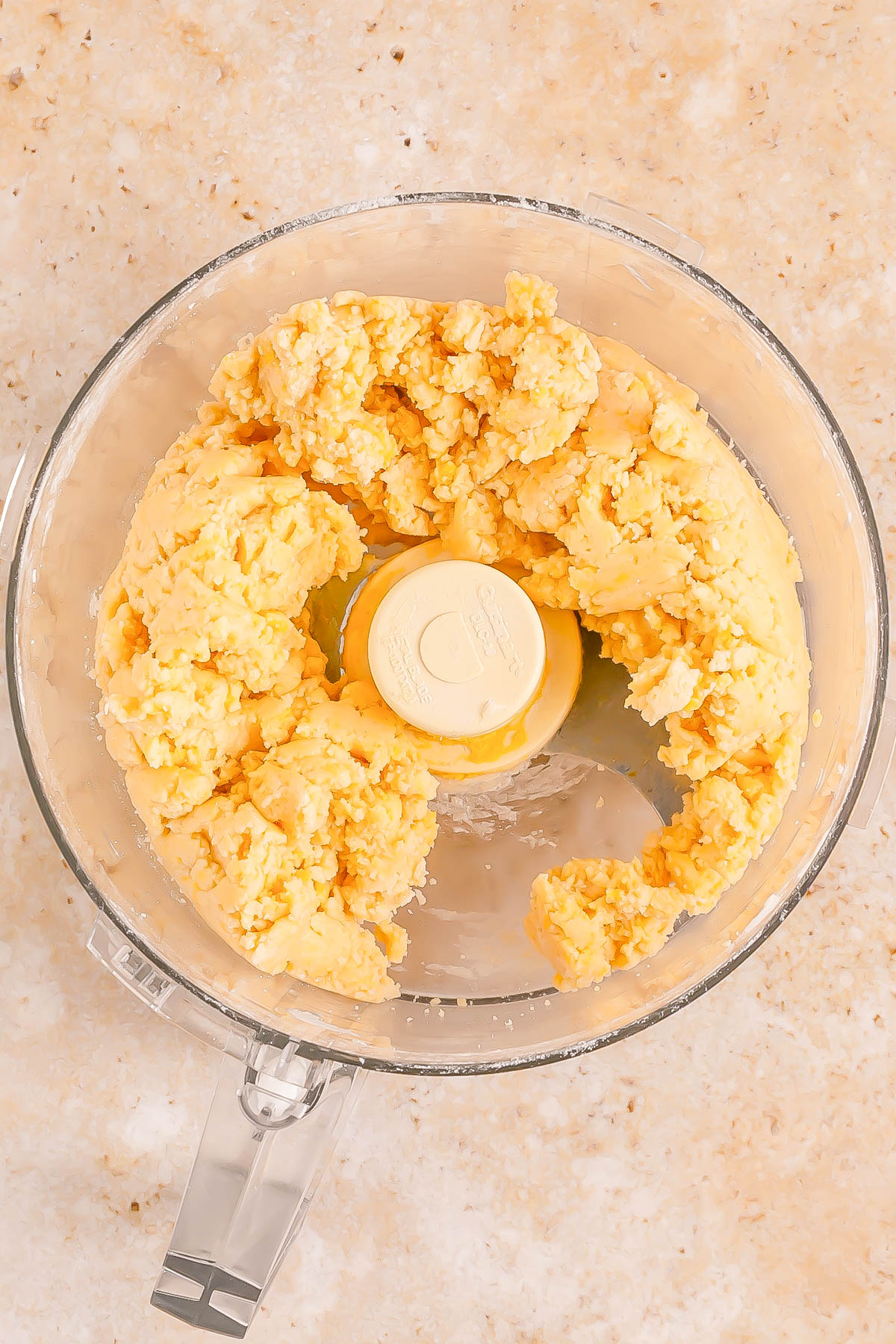 A food processor containing a ball of yellow dough sits on a light-colored countertop.