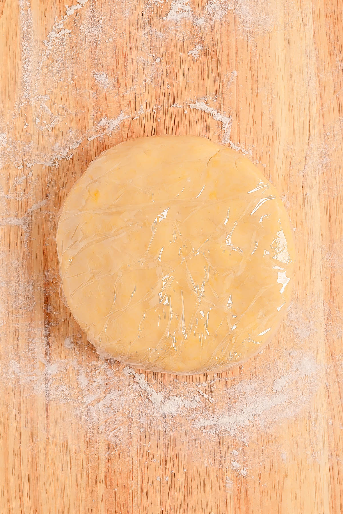 A round disc of dough wrapped in plastic wrap sits on a lightly floured wooden surface.