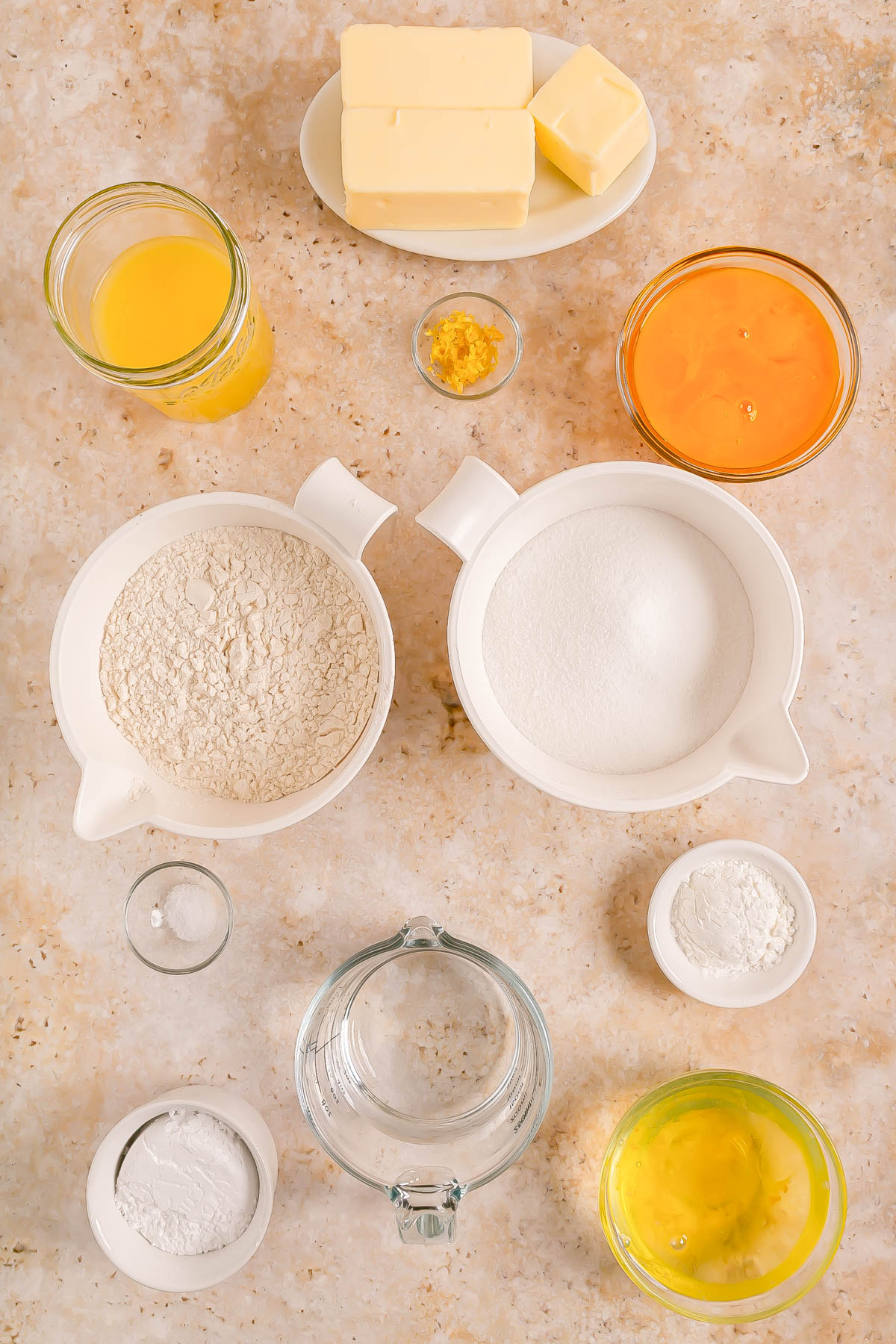 Various baking ingredients in separate bowls and containers, including flour, sugar, butter, eggs, orange juice, zest, baking powder, salt, and water, arranged on a countertop.