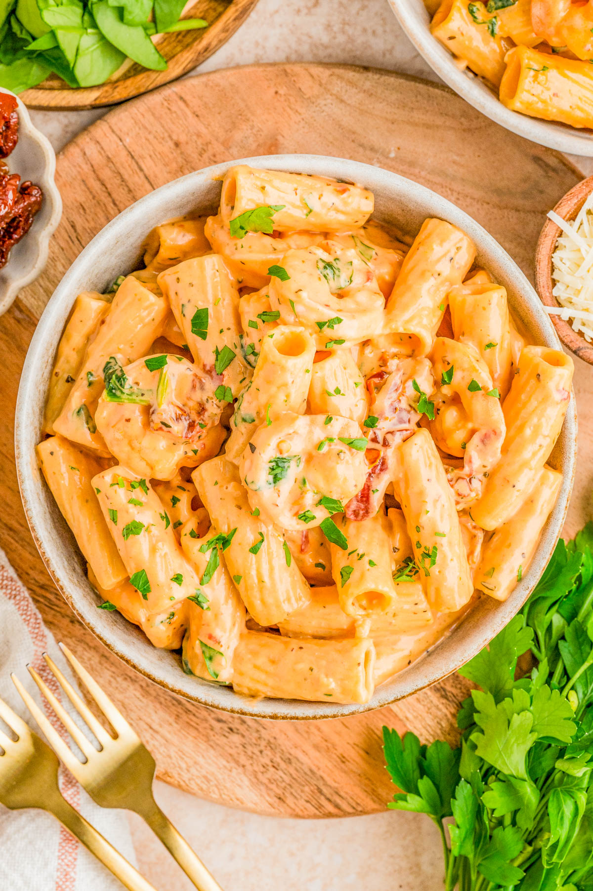 A bowl of creamy rigatoni pasta garnished with chopped parsley, placed on a wooden board with fresh greens, grated cheese, sun-dried tomatoes, and gold forks nearby.