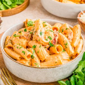 A bowl of creamy rigatoni pasta with shrimp, garnished with chopped parsley, sits on a wooden tray next to fresh herbs and a fork.
