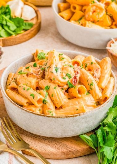A bowl of creamy rigatoni pasta with shrimp, garnished with chopped parsley, sits on a wooden tray next to fresh herbs and a fork.