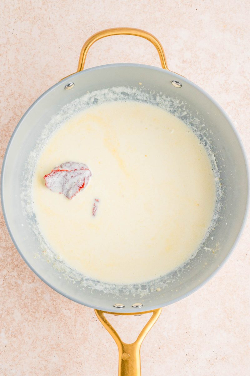 A saucepan with creamy white sauce and a partially submerged red onion half, shown on a light-colored surface.