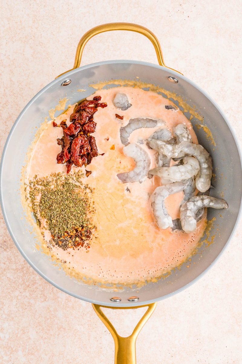 A pan contains raw shrimp, sun-dried tomatoes, dried herbs, and a creamy pink sauce base, ready to be cooked.