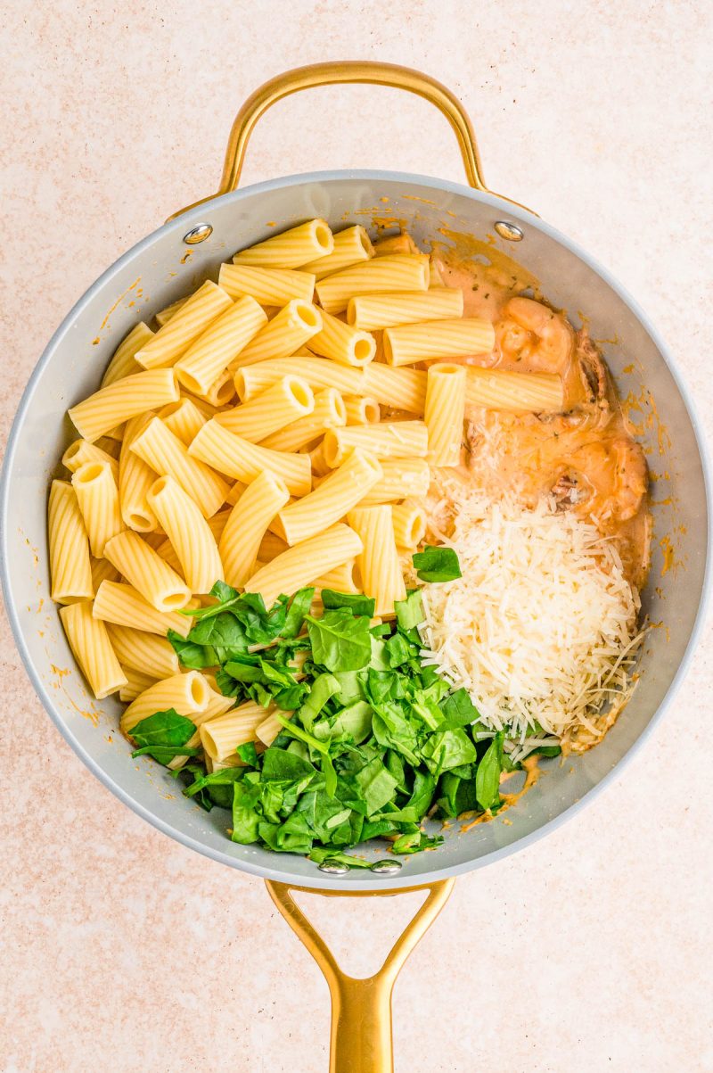 A pan containing cooked rigatoni pasta, chopped spinach, grated cheese, and creamy sauce, all separated before mixing.