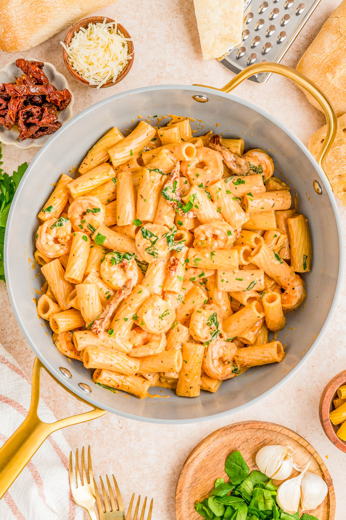 A skillet filled with creamy rigatoni pasta, shrimp, and chopped herbs, surrounded by grated cheese, sun-dried tomatoes, garlic, and fresh basil on a countertop.