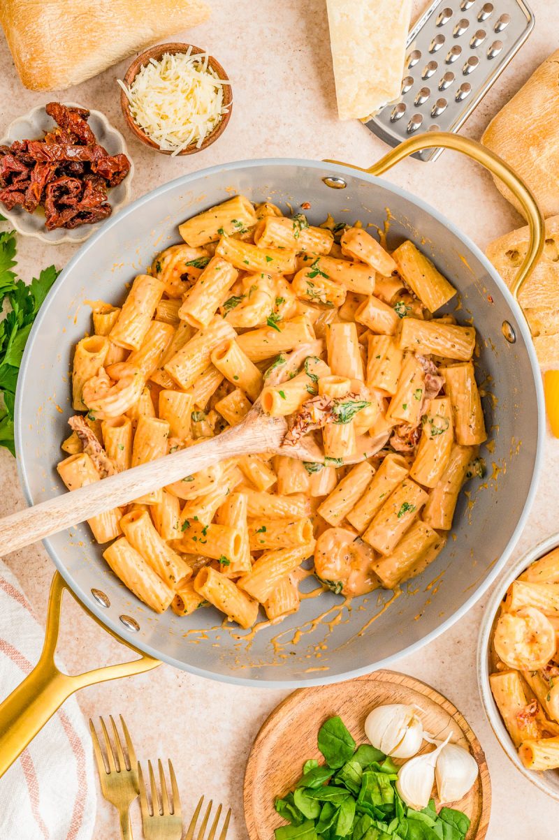 A skillet of creamy rigatoni pasta with shrimp and sun-dried tomatoes, garnished with herbs, surrounded by bread, cheese, garlic, spinach, and kitchen utensils.