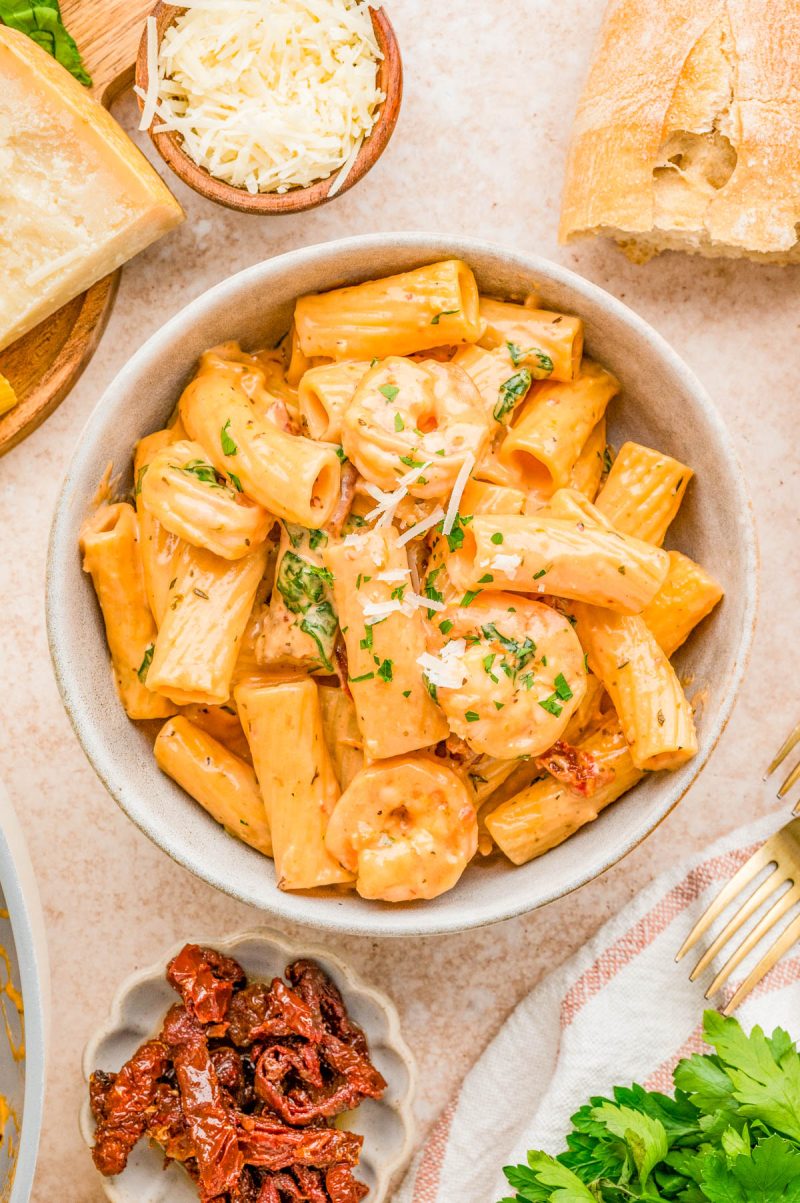 A bowl of creamy rigatoni pasta with shrimp, garnished with parsley and grated cheese. Surrounding items include bread, shaved cheese, sun-dried tomatoes, and fresh parsley.