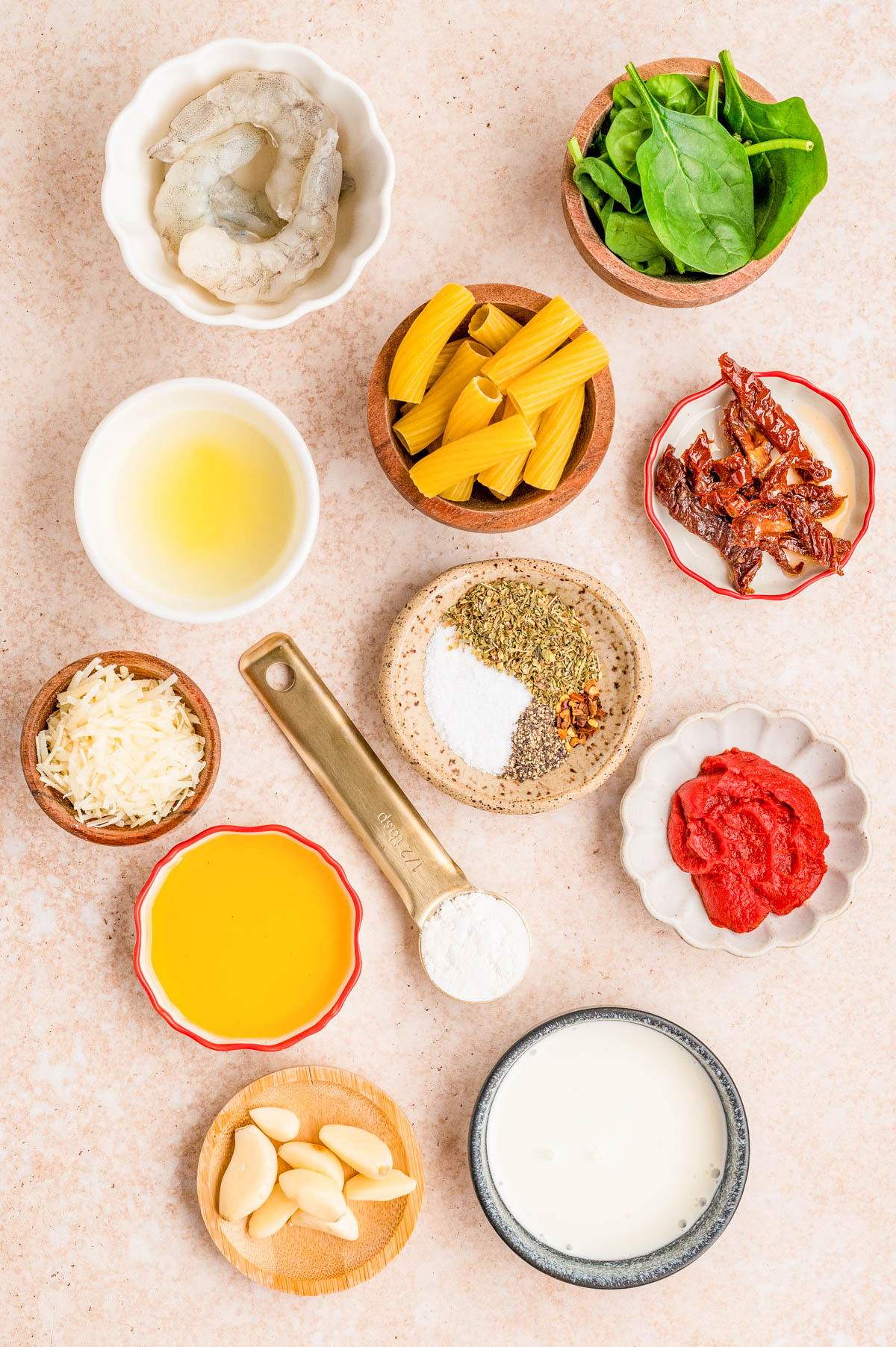 Top-down view of ingredients for a pasta dish, including shrimp, rigatoni, spinach, sun-dried tomatoes, cheese, spices, olive oil, flour, garlic, tomato paste, cream, and white wine.