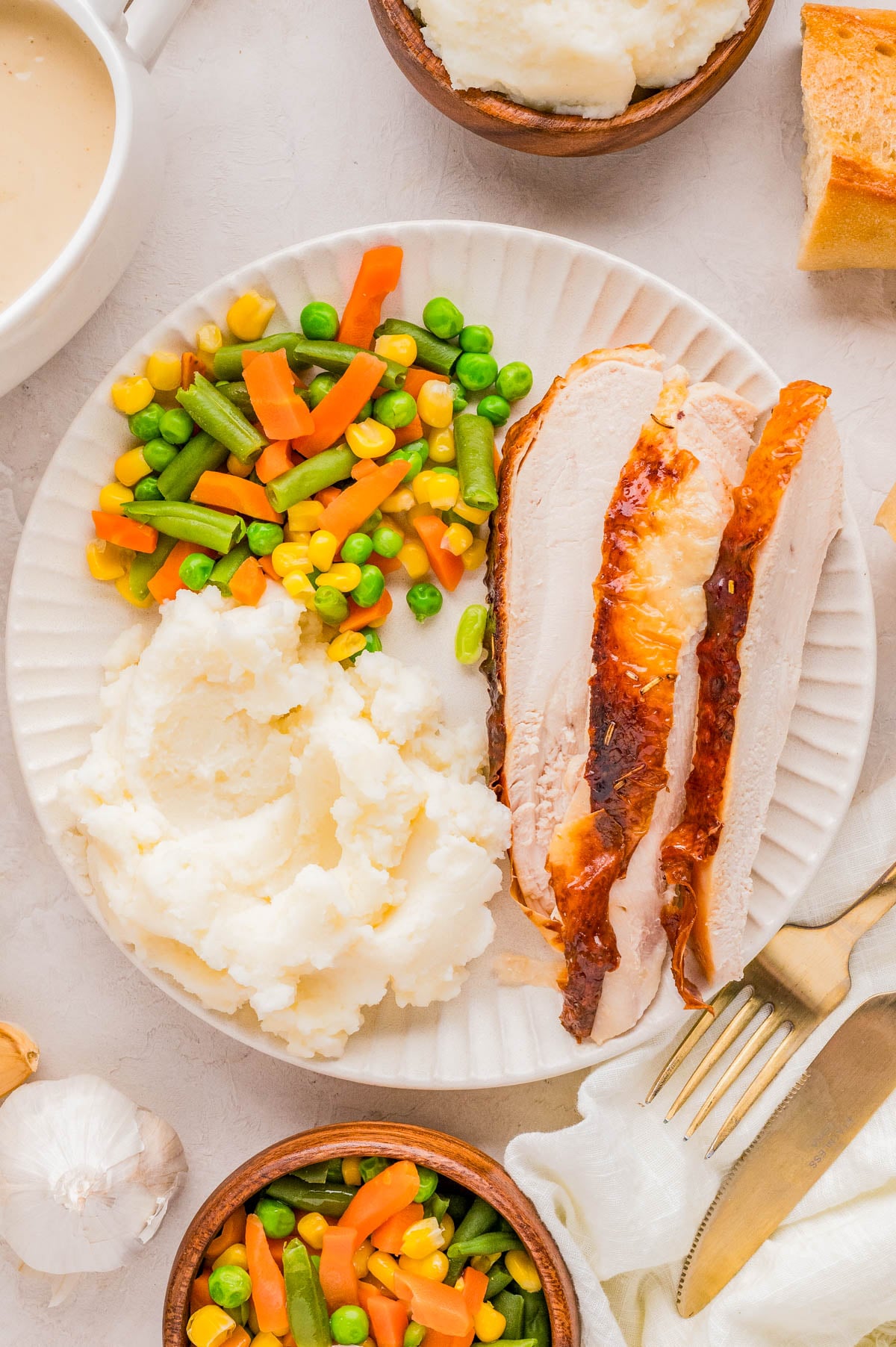 A plate with slices of roasted turkey, mashed potatoes, and mixed vegetables. Nearby are bowls of mashed potatoes and vegetables, a fork, knife, and bread.