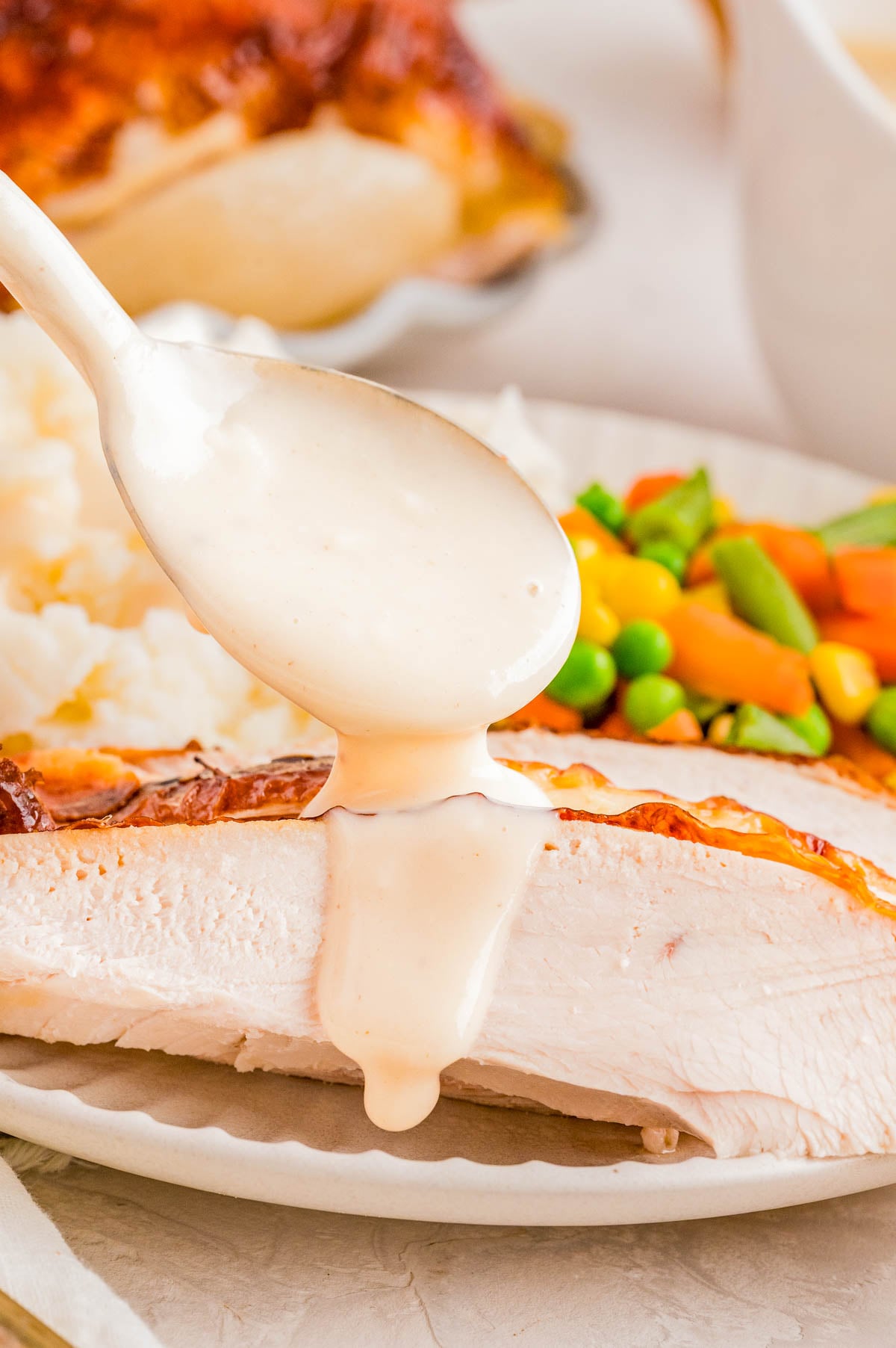 A spoon pours creamy gravy over a slice of turkey breast, with mashed potatoes and mixed vegetables on the plate.