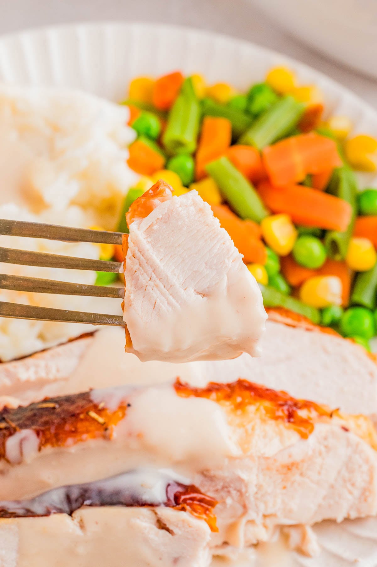 A fork holds a piece of turkey with gravy, with sliced turkey, mashed potatoes, and mixed vegetables on a plate in the background.