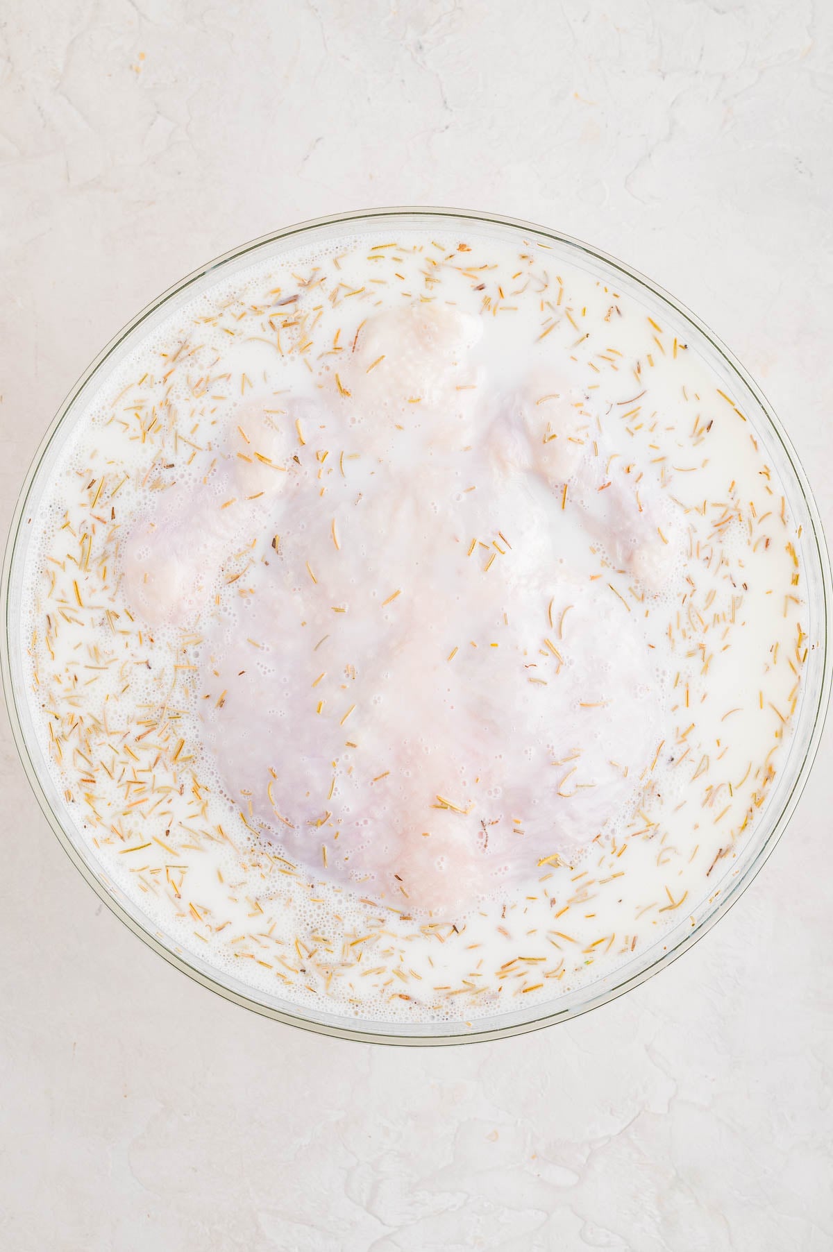 Raw whole chicken marinating in a bowl of milk with herbs sprinkled on top, set on a light-colored surface.