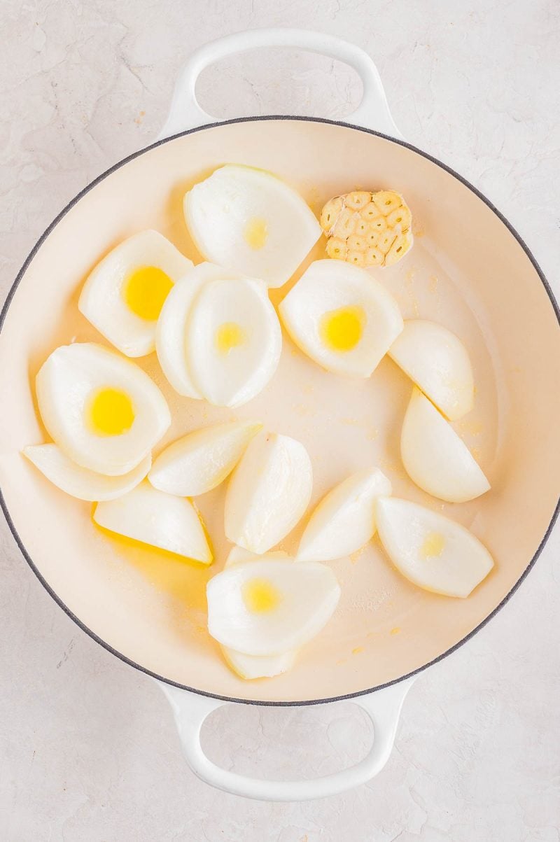 A white Dutch oven containing quartered onions, a halved head of garlic, and drizzles of oil, all arranged on a light surface.