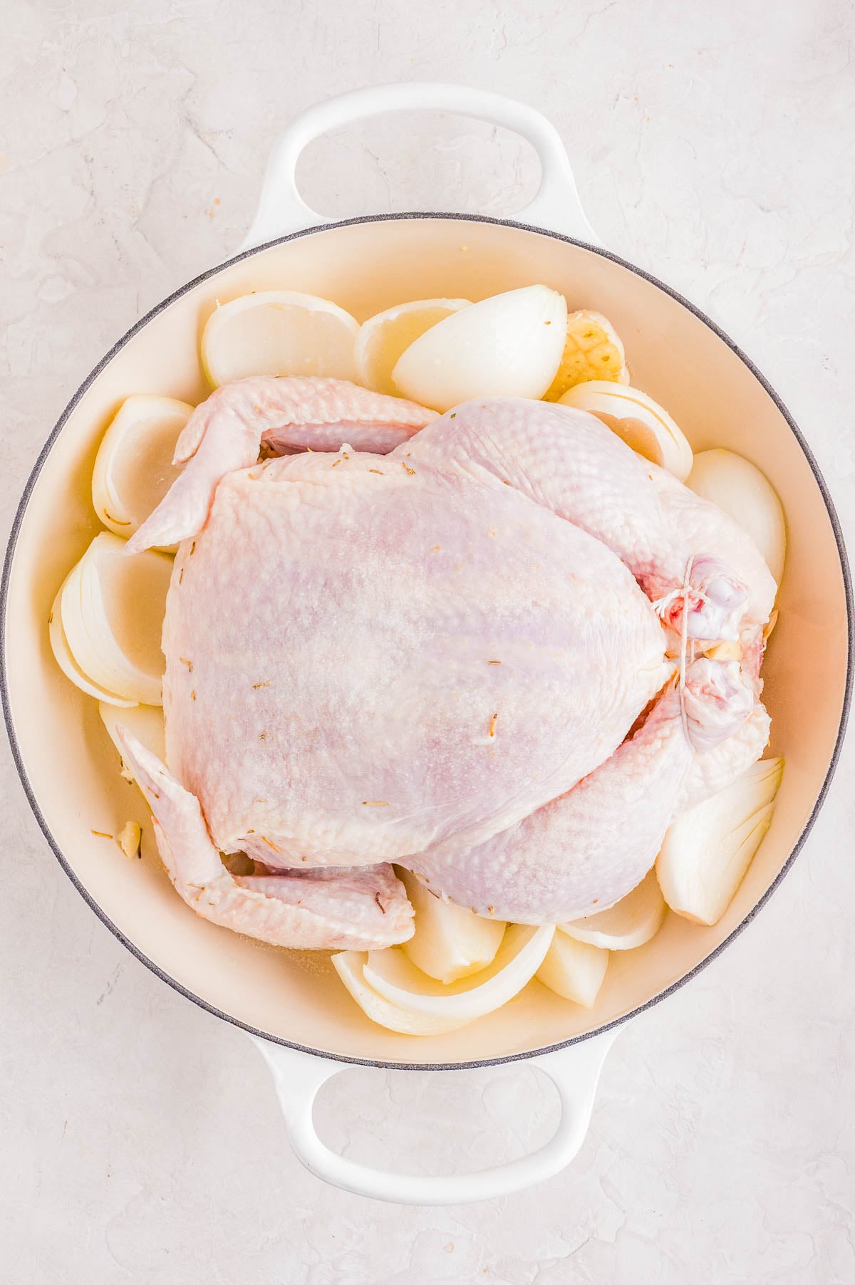 Whole raw chicken placed in a white pot surrounded by quartered onions on a light-colored surface.