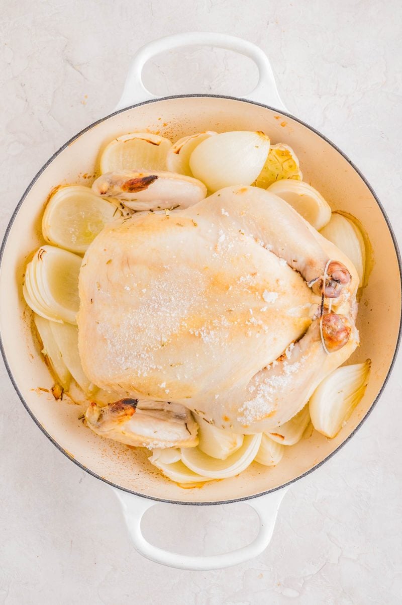 Whole raw chicken seasoned with salt, placed on top of sliced onions in a white Dutch oven, ready for roasting.