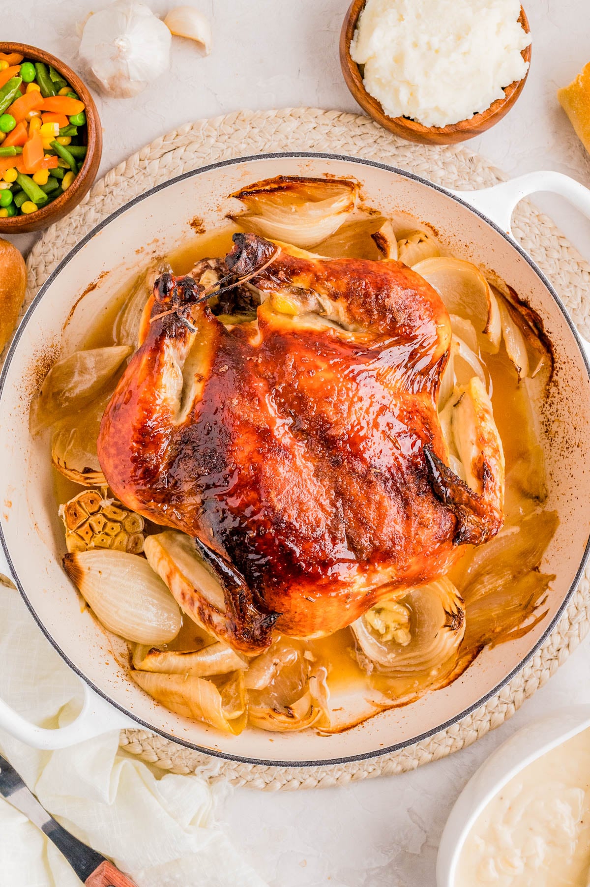 Roast chicken with crispy skin in a white pot, surrounded by cooked onions, with side dishes including mixed vegetables, mashed potatoes, and a bread roll nearby.