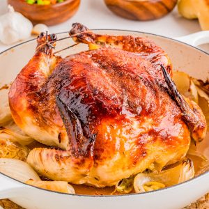 Whole roasted chicken in a white dish with browned skin, surrounded by onions, with bowls of mixed vegetables, rice, and rolls in the background.