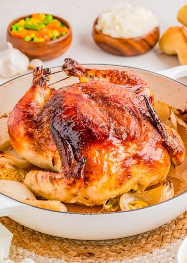 Whole roasted chicken in a white dish with browned skin, surrounded by onions, with bowls of mixed vegetables, rice, and rolls in the background.
