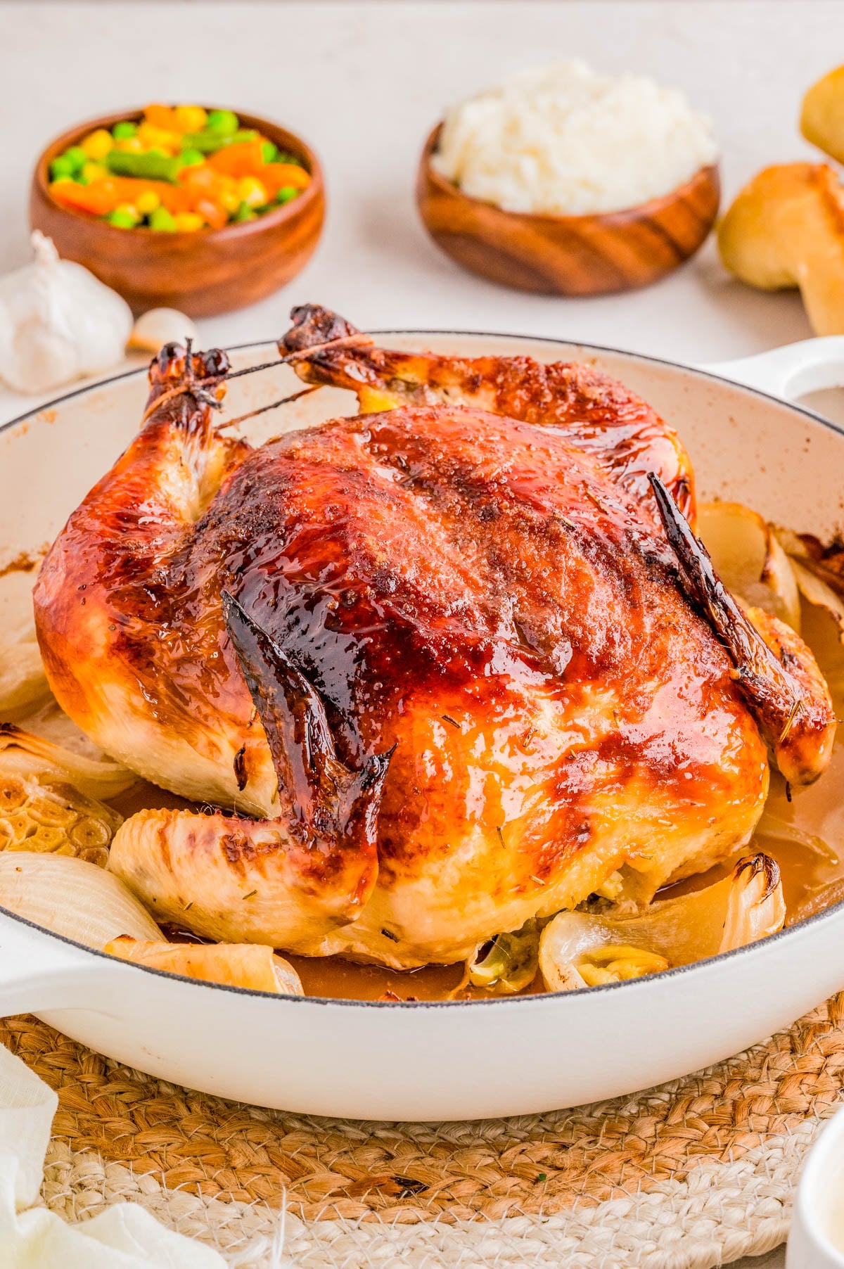 Whole roasted chicken in a white dish with browned skin, surrounded by onions, with bowls of mixed vegetables, rice, and rolls in the background.