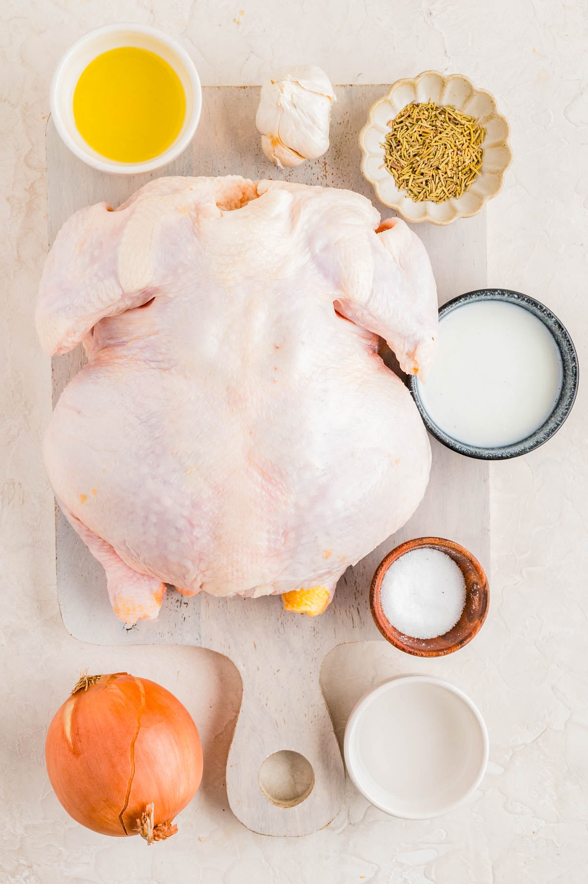 Raw whole chicken on a cutting board surrounded by olive oil, garlic, dried herbs, milk, salt, an onion, and a bowl of water.