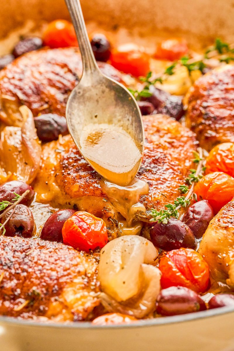 A spoon drizzling sauce over seasoned, cooked chicken thighs with cherry tomatoes, olives, shallots, and herbs in a pan.