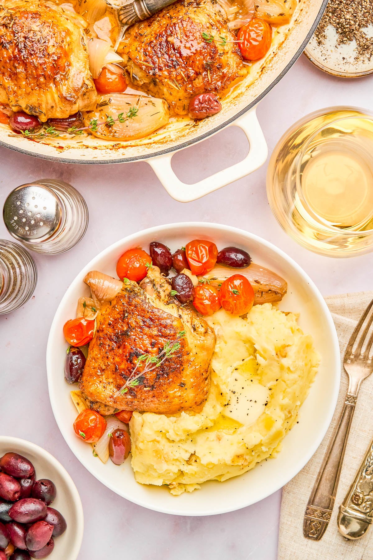 A plate of mashed potatoes with roasted chicken thigh, cherry tomatoes, shallots, olives, and a glass of white wine on the side.