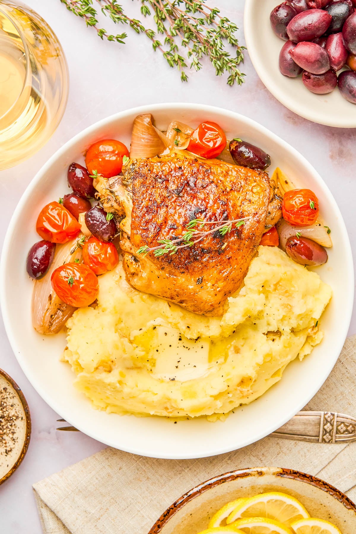 A plate of roasted chicken thigh on mashed potatoes, garnished with thyme, cherry tomatoes, olives, and shallots, with a glass of white wine and olives nearby.