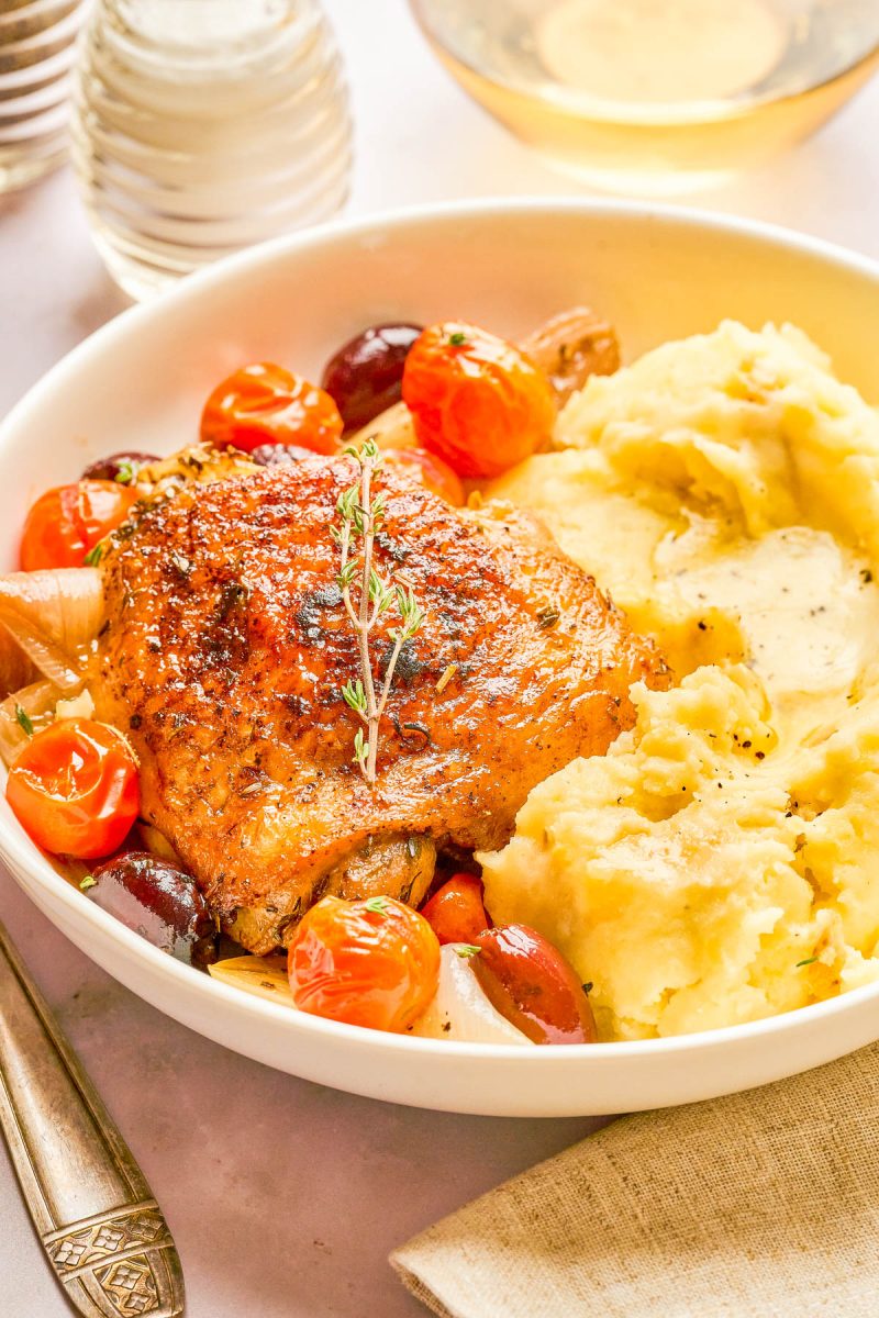 A plate with roasted chicken thigh, mashed potatoes, cherry tomatoes, olives, and a sprig of thyme. A fork and napkin are beside the dish.