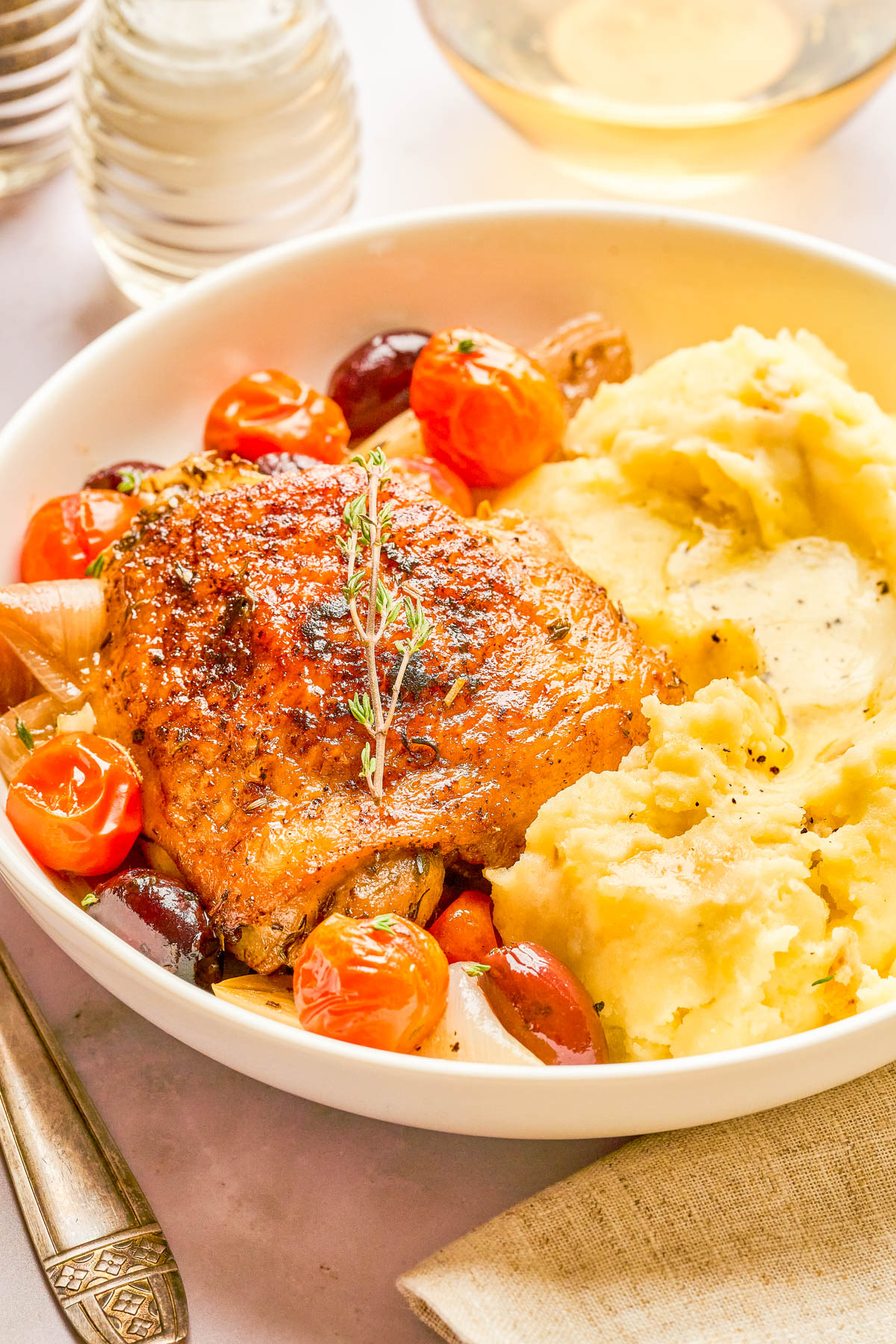 A plate with roasted chicken thigh, mashed potatoes, cherry tomatoes, olives, and a sprig of thyme. A fork and napkin are beside the dish.