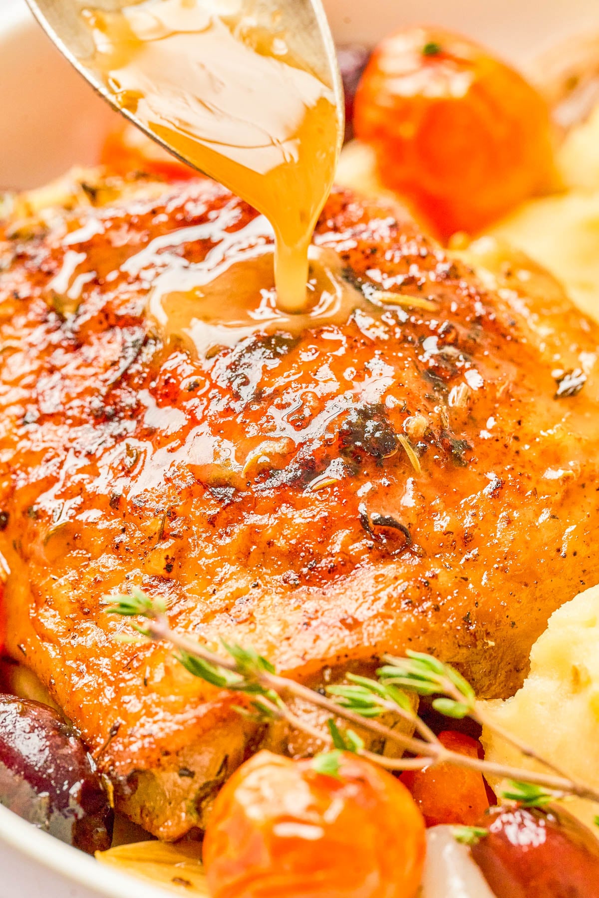 Close-up of a cooked, seasoned chicken thigh being drizzled with sauce, surrounded by vegetables and herbs.
