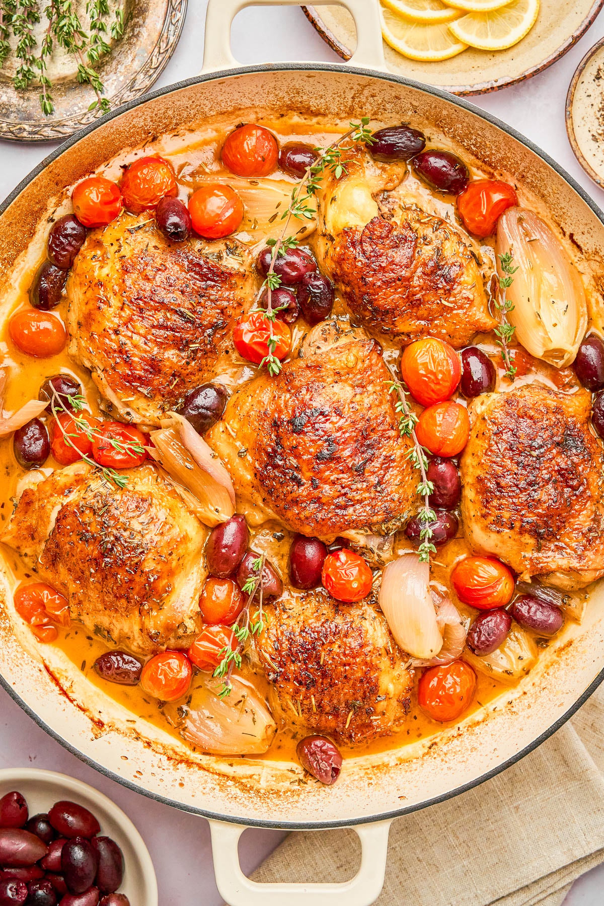 A skillet of roasted chicken thighs with crispy skin, cherry tomatoes, olives, shallots, and sprigs of thyme in a light sauce.