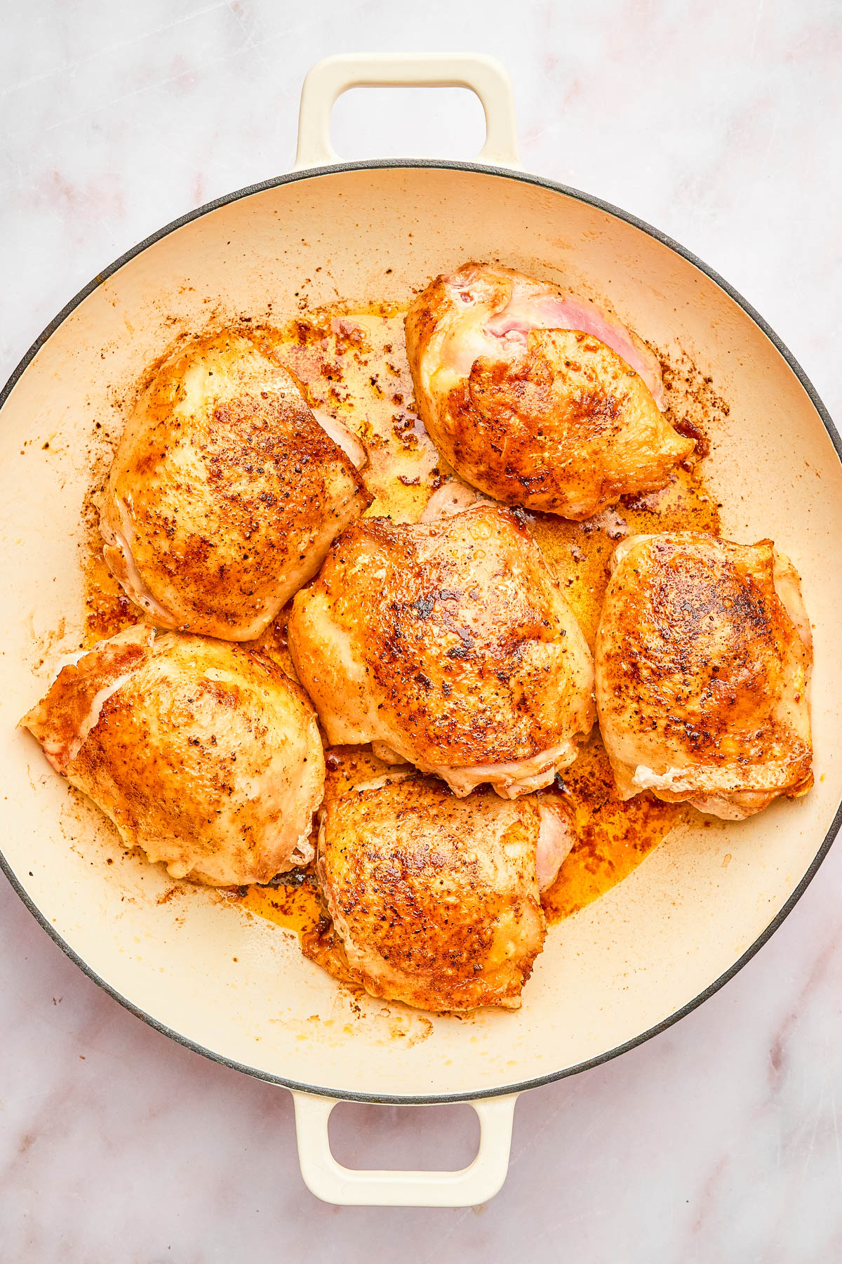 Six seasoned, cooked chicken thighs in a round, cream-colored skillet on a light surface.