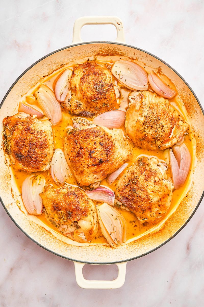 A round white pan filled with roasted chicken thighs and halved shallots in a golden sauce, viewed from above on a light surface.