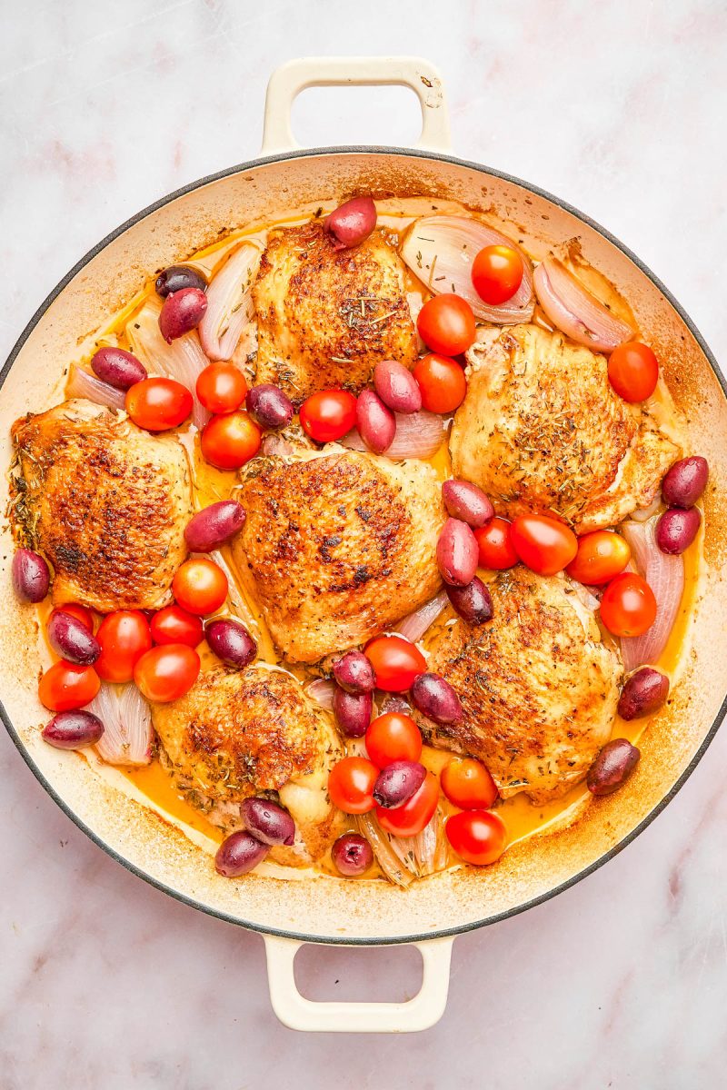 A skillet with roasted chicken thighs, cherry tomatoes, red grapes, shallots, and herbs in a light sauce.