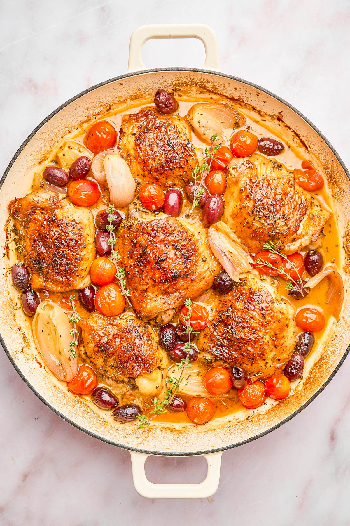 A skillet with roasted chicken thighs, cherry tomatoes, shallots, olives, and sprigs of thyme in a savory sauce.