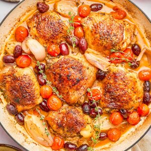A skillet with roasted chicken thighs, cherry tomatoes, shallots, olives, and herbs in a savory sauce.